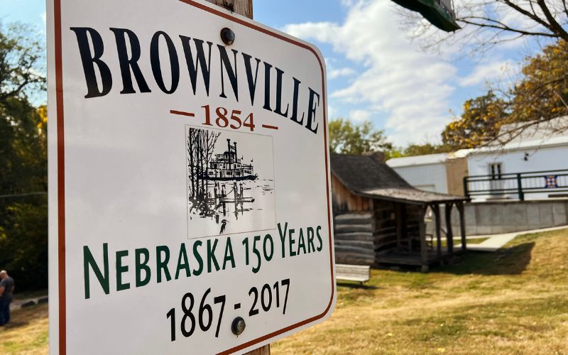 A sign for Brownville inn Boettner Park with the 1854 Didier Log Cabin in the background.