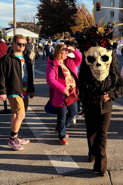 A person in black wearing a giant skull with flowers on top walks among the crowd of festival goers during the Day of the Dead