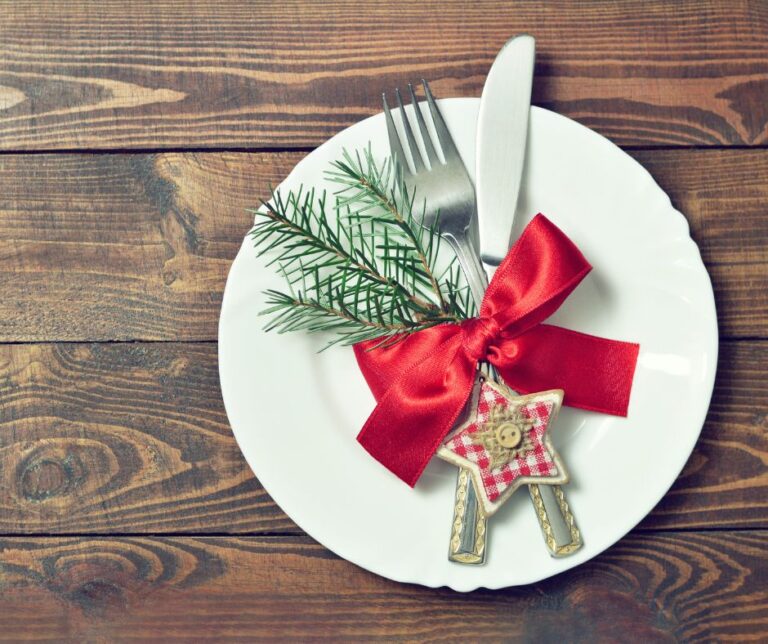 A plate with a knife and forked wrapped decoratively with a red ribbon, holly sprig, and ornament.