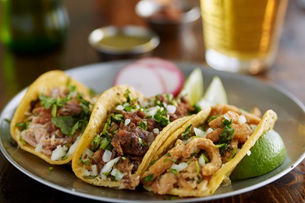 Three street style tacos on a silver plate with a glass of beer in the background.