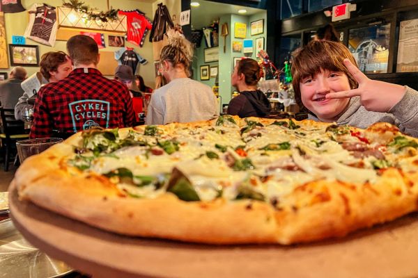 A kid shows a peace symbol from behind a large pizza at The Pedaler's Pub, a bike-themed pizza shop in Bentonville