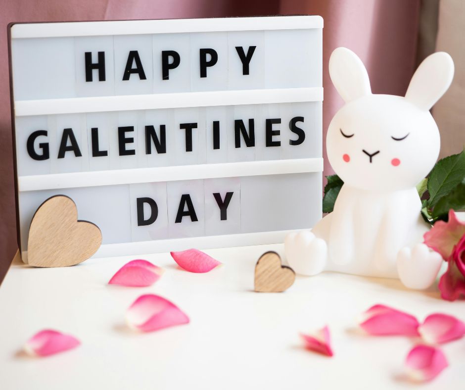 A sign reads "Happy Galentine's Day" in front of some flower petals and wooden hearts. There's a cute white bunny statue next to it.